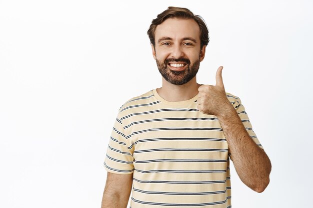 Lächelnder bärtiger Mann, der zustimmend Daumen nach oben zeigt und zustimmt, im T-Shirt auf weißem Hintergrund zu stehen