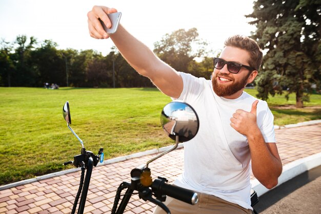 Lächelnder bärtiger Mann in der Sonnenbrille, die draußen auf modernem Motorrad sitzt und Selfie auf seinem Smartphone macht