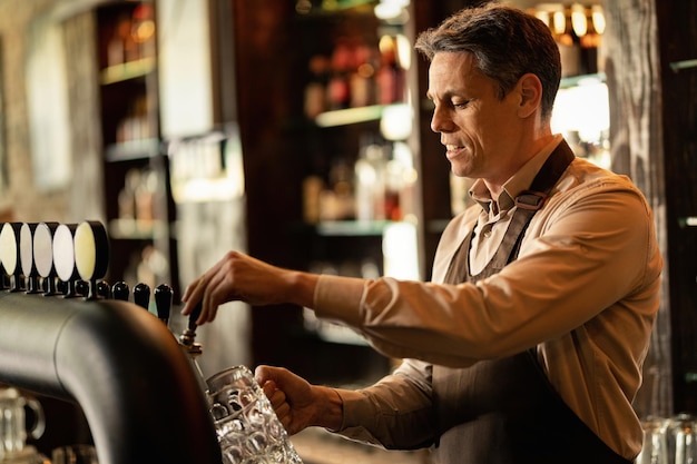 Kostenloses Foto lächelnder barkeeper, der in einer kneipe arbeitet und bier aus einem zapfhahn gießt