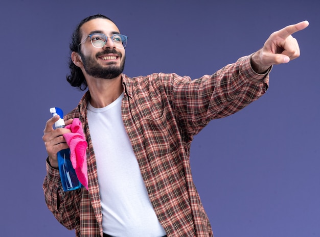 Lächelnder Blick auf den jungen, gutaussehenden Putzmann der Seite, der das T-Shirt hält, das Sprühflasche mit Lappenpunkten an der Seite hält, die auf blauer Wand lokalisiert wird