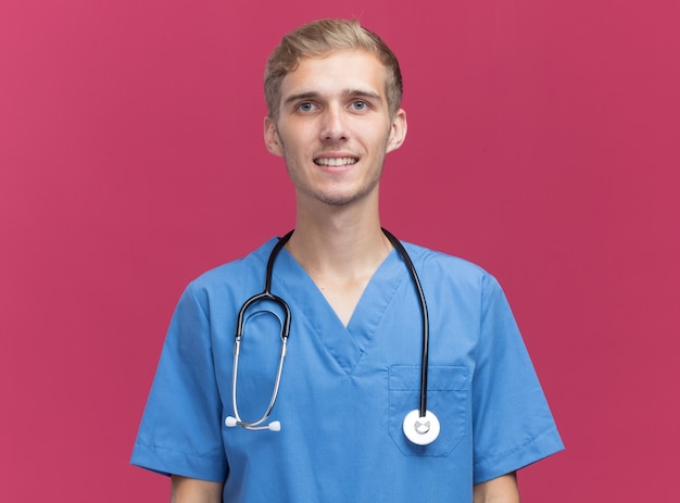 Lächelnder Blick auf den jungen männlichen Arzt der Kamera, der Arztuniform mit Stethoskop lokalisiert auf rosa Wand mit Kopienraum trägt