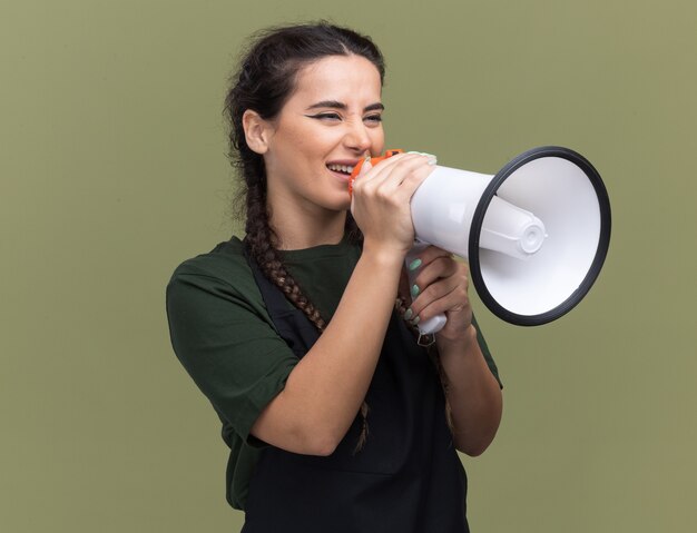 Lächelnder Blick auf die Seite junger weiblicher Friseur in Uniform spricht auf Lautsprecher isoliert auf olivgrüner Wand