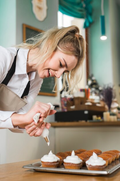 Lächelnder blonder weiblicher Bäcker, der mit Sahne kleine Kuchen verziert