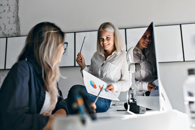 Lächelnder blonder weiblicher Manager, der Infografik und Bleistift hält, während auf Tisch sitzt. Innenporträt von zwei Frauen, die mit Computer im Büro arbeiten.