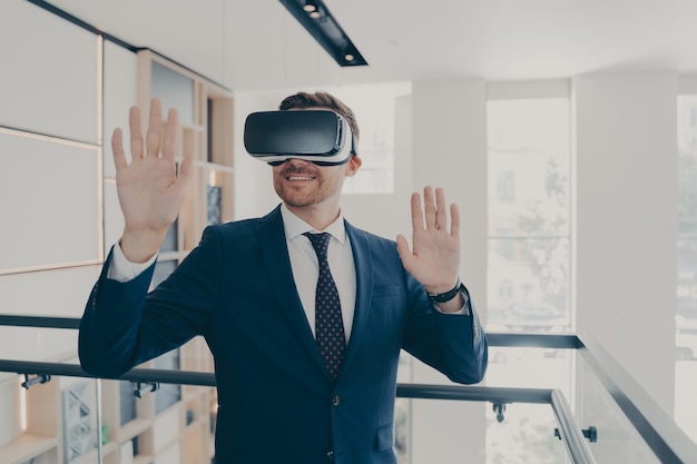 Lächelnder büroangestellter im anzug mit vr-headset, der luft berührt und während der arbeit in der virtuellen realität gestikuliert, geschäftsmann, der ein geschäftsprojekt mit 3d-brille verwaltet, während er im bürokorridor steht