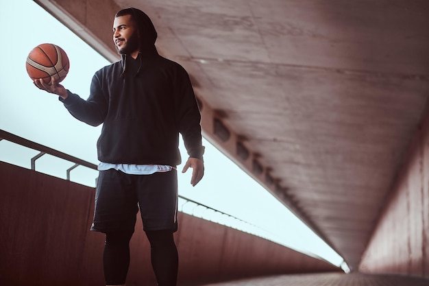 Lächelnder dunkelhäutiger Typ in schwarzem Hoodie und Sportshorts hält einen Basketball, während er auf einem Fußweg unter einer Brücke steht.