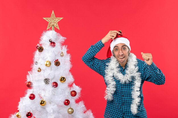 Lächelnder emotionaler lustiger junger Mann mit Weihnachtsmannhut in einem blauen gestreiften Hemd