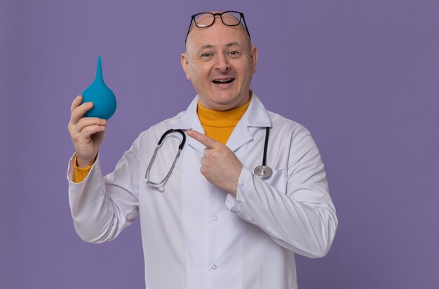 Lächelnder erwachsener Mann mit Brille in Arztuniform mit Stethoskop, das Einlauf hält und betrachtet