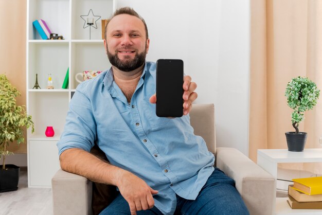 Lächelnder erwachsener slawischer Mann sitzt auf Sessel, der Telefon innerhalb des Wohnzimmers hält