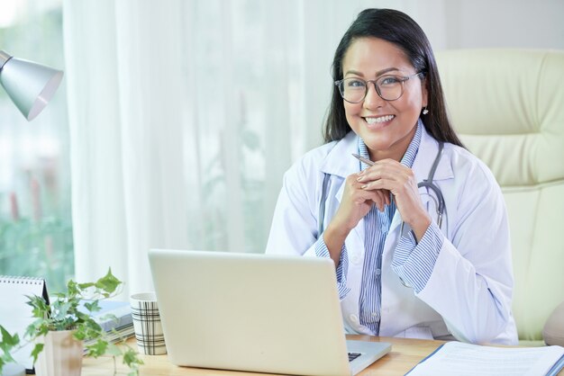 Lächelnder ethnischer Doktor, der am Schreibtisch im Büro sitzt