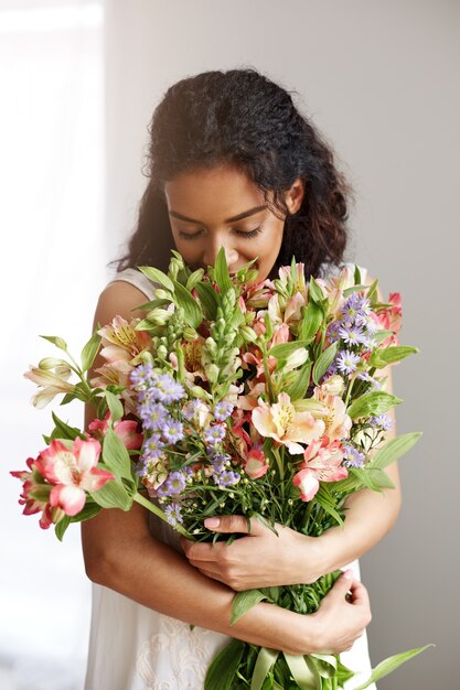 Lächelnder Florist der afrikanischen Frau, der Schnüffelstrauß von Alstroemerias über weißer Wand hält. Geschlossene Augen.