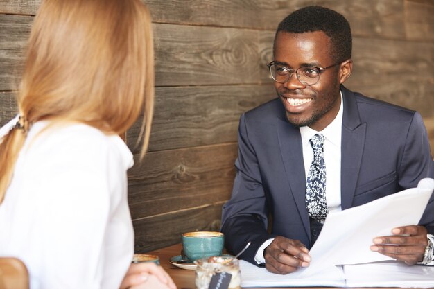 Lächelnder fröhlicher afroamerikanischer Unternehmer, der Brille und formellen Anzug trägt