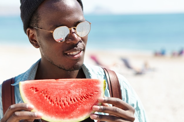 Lächelnder fröhlicher junger schwarzer Hipster, der große Scheibe reife und saftige Wassermelone hält, die vorwegnehmenden Blick hat