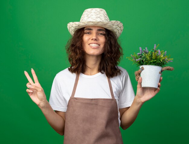 Lächelnder Gärtner der jungen Frau in der Uniform, die Gartenhut hält, der Blume im Blumentopf hält, die Friedensgeste lokalisiert auf Grün zeigt