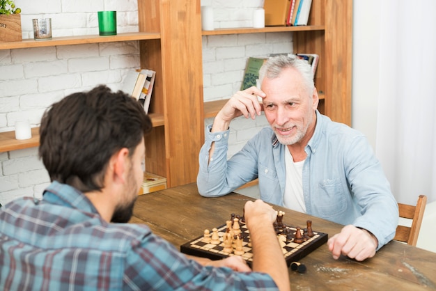 Lächelnder gealterter Mann und junger Kerl, die bei Tisch Schach nahe Bücherregalen spielen