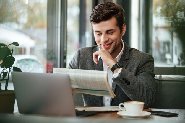 Lächelnder Geschäftsmann, der durch die Tabelle im Café mit Laptop-Computer beim Ablesen der Zeitung sitzt