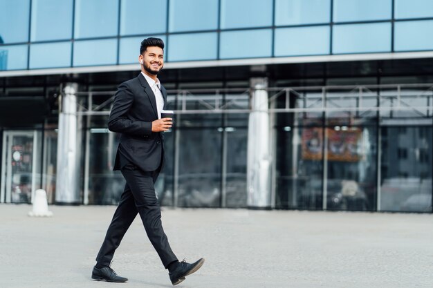 Lächelnder glücklicher Inder geht im Büro zur Arbeit, hinter ihm ein modernes Gebäude