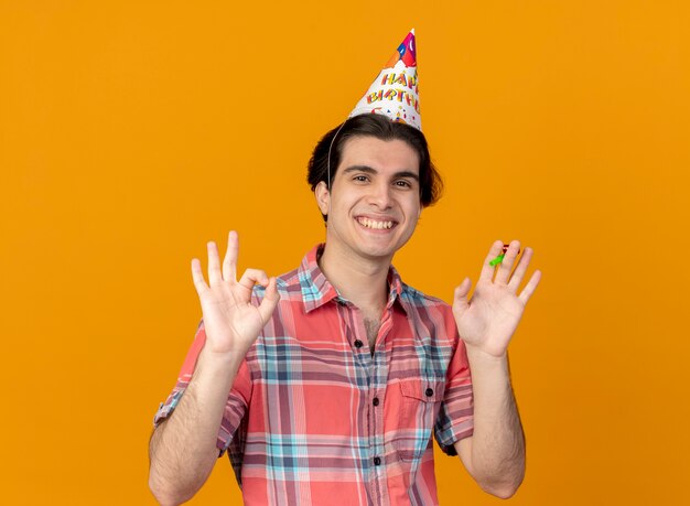 Lächelnder gutaussehender kaukasischer Mann mit Geburtstagsmütze gestikuliert ok Handzeichen und hält Partypfeife