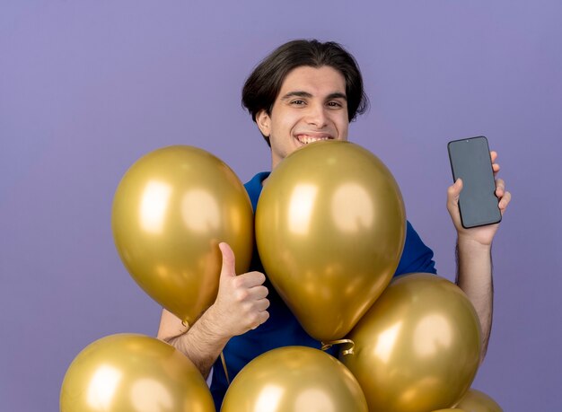 Lächelnder gutaussehender kaukasischer Mann steht mit Heliumballons Daumen hoch und hält Telefon