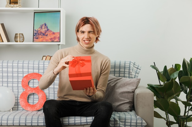 Lächelnder, gutaussehender Kerl am glücklichen Frauentag, der das Geschenk auf dem Sofa im Wohnzimmer hält