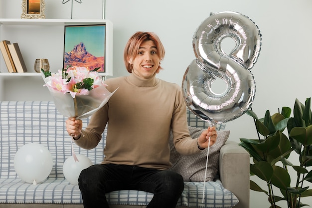 Lächelnder gutaussehender Kerl am glücklichen Frauentag, der den Ballon Nummer acht und den Blumenstrauß auf dem Sofa im Wohnzimmer hält