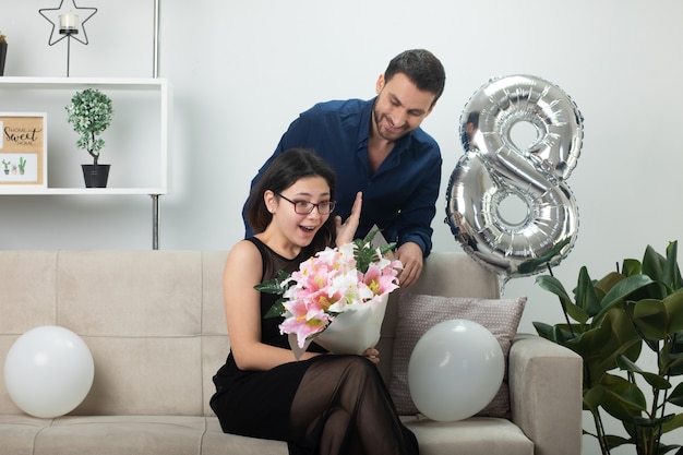Lächelnder gutaussehender Mann, der aufgeregte hübsche junge Frau in optischer Brille mit Blumenstrauß ansieht, die am internationalen Frauentag im März auf der Couch im Wohnzimmer sitzt