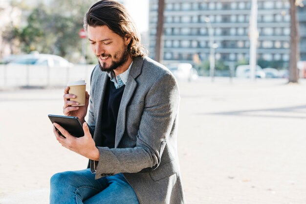 Lächelnder gutaussehender Mann, der den Handy hält Wegwerfkaffeetasse betrachtet