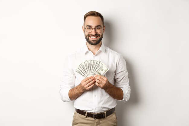 Lächelnder gutaussehender Mann, der Geld hält, Dollar zeigt, stehend