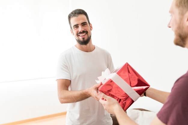 Lächelnder gutaussehender Mann, der Rot eingewickelte Geschenkbox zu seinem Freund nimmt