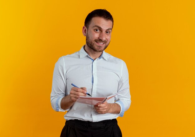 Lächelnder gutaussehender Mann hält Stift und Notizbuch, die auf orange Wand lokalisiert suchen