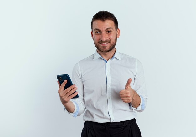 Lächelnder gutaussehender Mann hält Telefon und Daumen isoliert auf weißer Wand