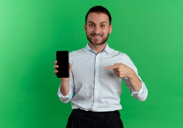 Lächelnder gutaussehender Mann hält und zeigt auf Telefon lokalisiert auf grüner Wand