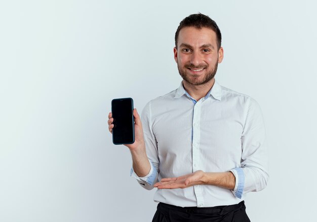 Lächelnder gutaussehender Mann hält und zeigt auf Telefon mit Hand lokalisiert auf weißer Wand