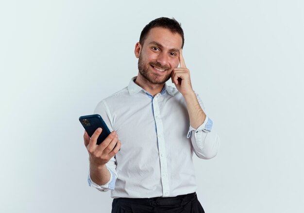 Lächelnder gutaussehender Mann legt Finger auf Tempel, der Telefon lokalisiert auf weißer Wand hält