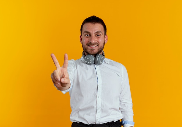 Lächelnder gutaussehender Mann mit Kopfhörern am Hals gestikuliert Siegeshandzeichen lokalisiert auf orange Wand
