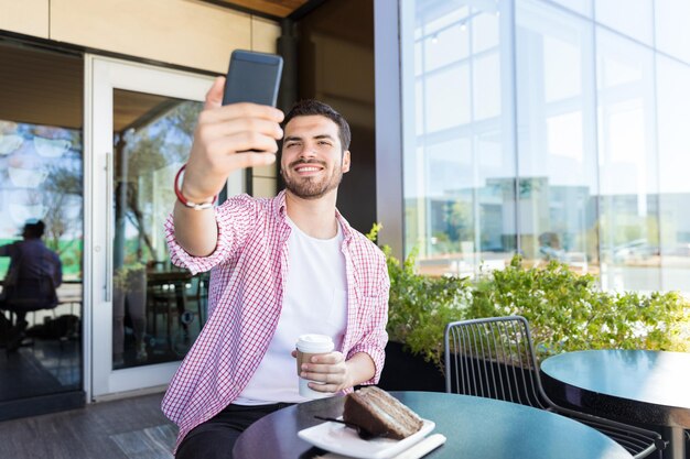 Lächelnder hispanischer Influencer, der ein Selfie macht, während er Dessert und Kaffee im Café genießt