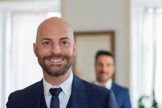 Lächelnder homosexueller Mann im offiziellen Anzug, der in die Kamera schaut. Nahaufnahme eines glücklichen Schwulen, der sich für die Hochzeitszeremonie anzieht. Stehend im Hotelzimmer mit seinem Partner im Hintergrund. Liebe, Emotionskonzept