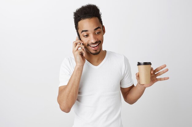 Lächelnder hübscher Afroamerikanermann, der am Telefon spricht und Kaffee mit glücklichem Ausdruck trinkt