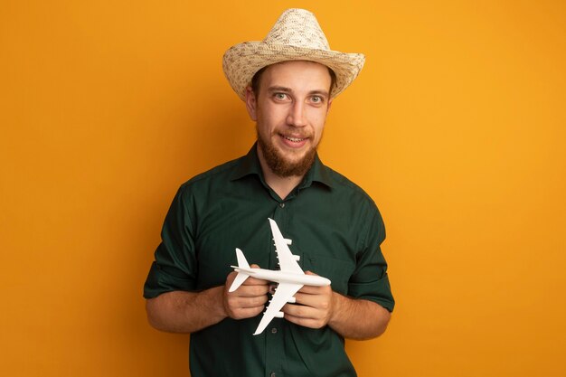 Lächelnder hübscher blonder Mann mit Strandhut, der Modellflugzeug auf Orange hält