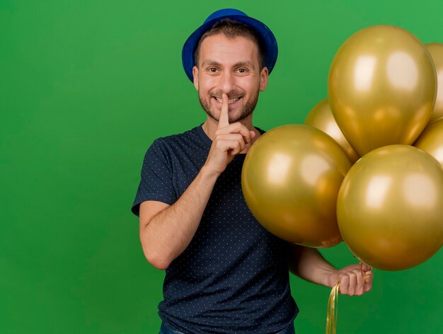 Lächelnder hübscher kaukasischer Mann, der blauen Parteihut trägt, gestikuliert Stille und hält Heliumballons lokalisiert auf grünem Hintergrund mit Kopienraum