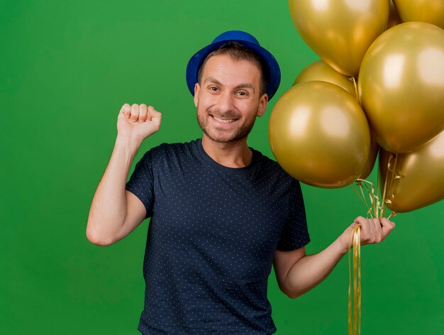 Lächelnder hübscher kaukasischer Mann, der blauen Parteihut trägt, hält Heliumballons und hält Faust lokalisiert auf grünem Hintergrund mit Kopienraum