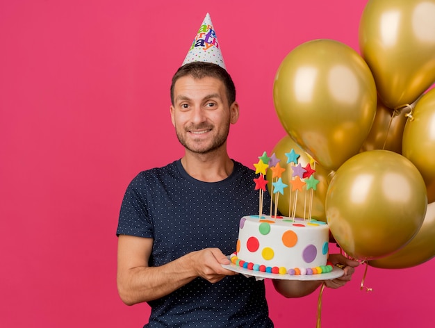Lächelnder hübscher kaukasischer Mann, der Geburtstagskappe trägt, hält Geburtstagstorte und Heliumballons lokalisiert auf rosa Hintergrund mit Kopienraum