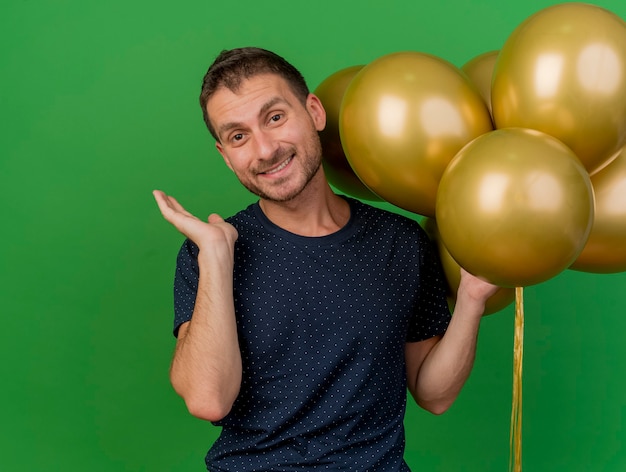 Lächelnder hübscher kaukasischer Mann steht mit erhabener Hand und hält Heliumballons lokalisiert auf grünem Hintergrund mit Kopienraum
