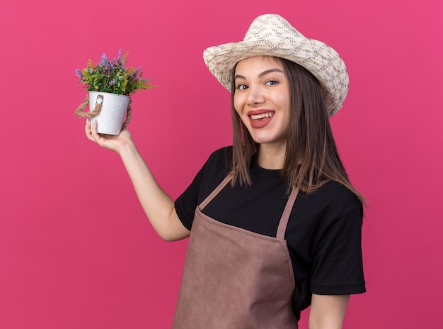 Lächelnder hübscher kaukasischer weiblicher Gärtner, der Gartenhut hält, der Blumentopf lokalisiert auf rosa Wand mit Kopienraum hält