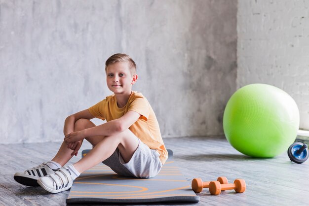 Lächelnder Junge, der auf Übungsmatte mit Dummkopf sitzt; pilates ball und roller slide