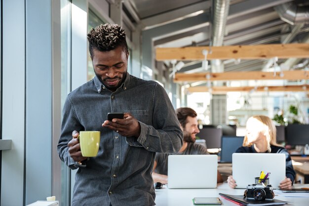 Lächelnder junger afrikanischer Mann, der im Büro steht und per Telefon plaudert