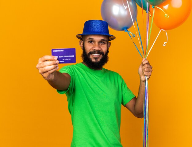 Lächelnder junger Afroamerikaner mit Partyhut, der Luftballons hält und Kreditkarte in die Kamera hält