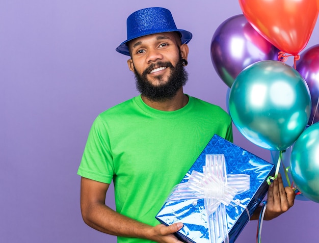 Lächelnder junger Afroamerikaner mit Partyhut, der Luftballons mit Geschenkbox hält