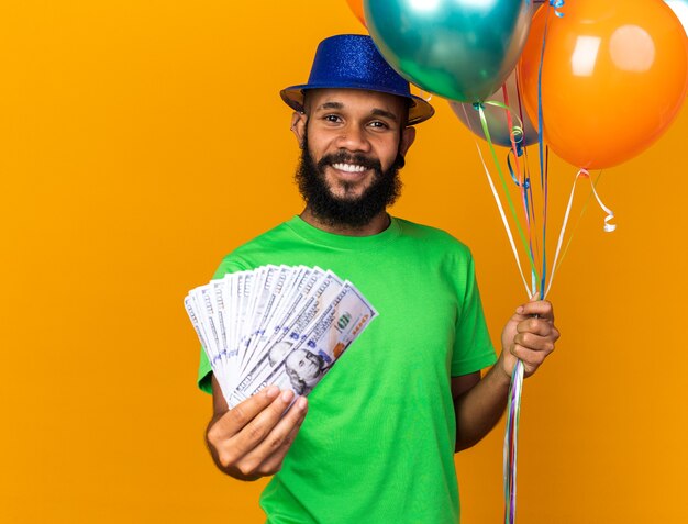 Lächelnder junger Afroamerikaner mit Partyhut mit Luftballons und Bargeld