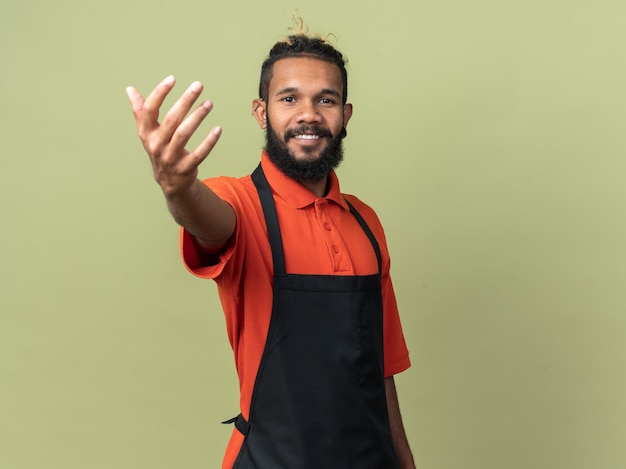 Lächelnder junger afroamerikanischer Friseur in Uniform, der die Hand in Richtung ausstreckt